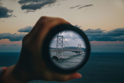 Cropped hand of person holding lens with reflection of bridge against sky