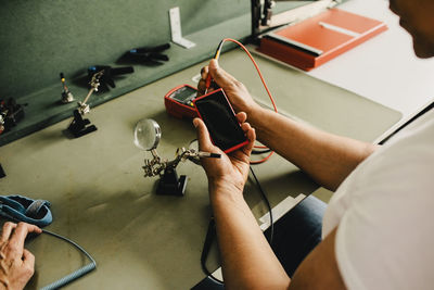 High angle view of male technician examining smart phone using multimeter at repair shop