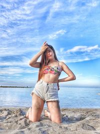 Full length portrait of young woman kneeling at beach against sky