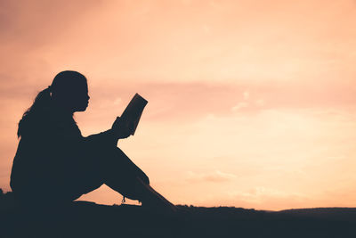 Silhouette young woman reading book while sitting on field against sky during sunset