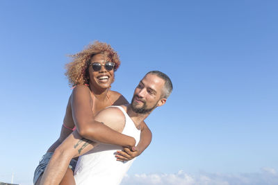 Low angle view of man piggybacking cheerful girlfriend against blue sky