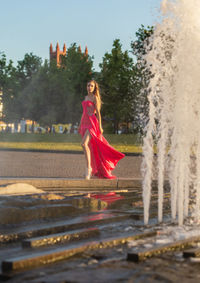 Young woman in pink evening gown standing by fountain in park