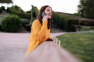 Happy caucasian woman wearing yellow pullover talking on mobile phone outdoors in park 