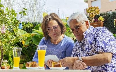 Senior woman showing digital tablet to man while having breakfast while sitting in yard