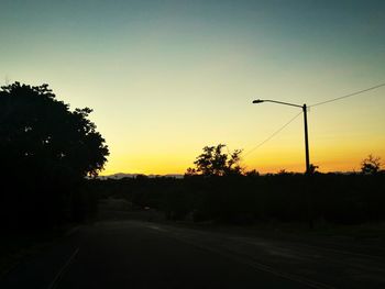 Road by silhouette trees against clear sky at sunset