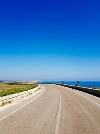 Empty road by sea against clear blue sky