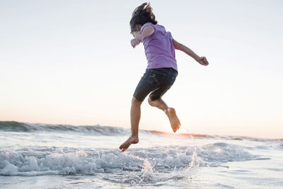 Low angle view of girl jumping on waves at beach against clear sky during sunset