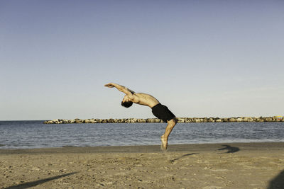 Full length of shirtless man jumping at beach against clear sky