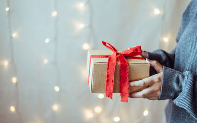 Midsection of woman holding christmas gift against illuminated wall at home