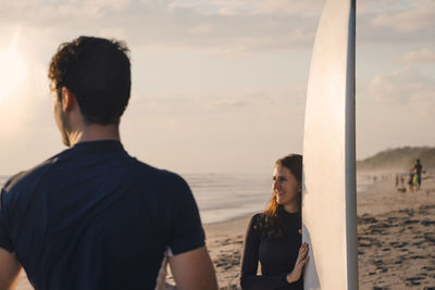 Young man with female friend holding surfboard at beach during sunset