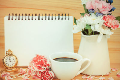 Close-up of coffee cup with flower vase and spiral notebook against wooden wall