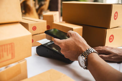 Cropped hands of man using mobile phone and card reader by boxes on table