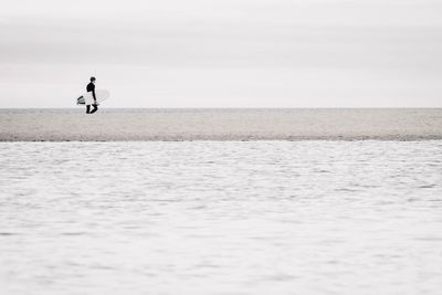 Distant view of man holding surfboard at beach against sky