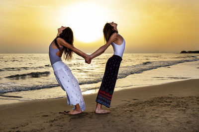 Two young women back to back hold their hands and stretch their back on the beach at sunset. 