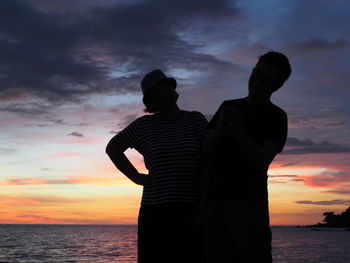 Silhouette man and woman standing by sea against sky during sunset