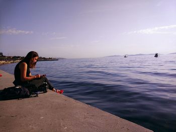 Side view of young woman sitting on sea against sky