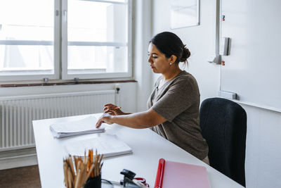 Focused female teacher checking test papers while sitting near table in classroom at junior high school