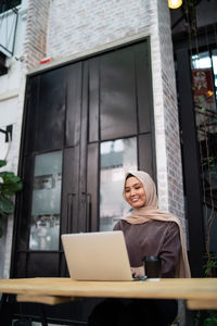 Young woman wearing hijab using laptop on table while sitting at sidewalk cafe
