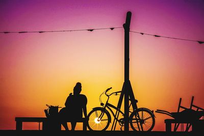 Silhouette people riding bicycle on street against orange sky