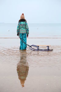 Reflection of girl with sledge standing at beach during winter