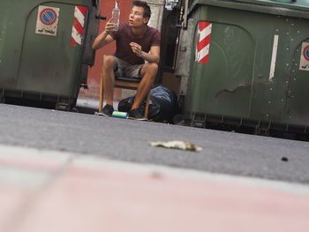 Surprised young man holding alcohol bottle amidst garbage cans