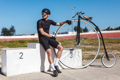 Low angle of serious thoughtful man in black sportswear and helmet looking away and thinking about future competition while sitting alone on champion podium and leaning on retro high wheel bicycle against blurred sports stadium