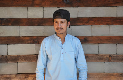 A young guy wearing cap backwards, looking sideways while standing against wooden and stone wall