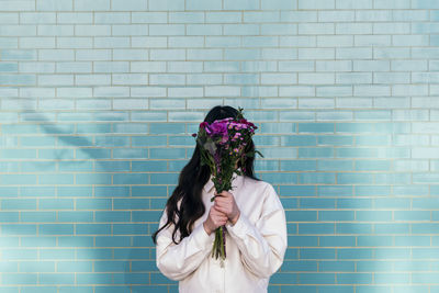 Young woman hiding face behind bouquet in front of brick wall