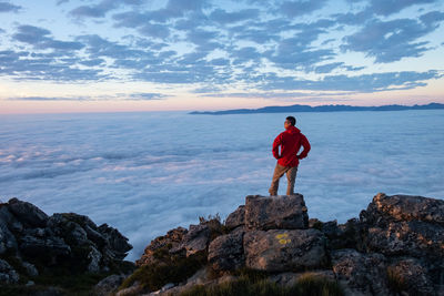 Man in red jacket  standing on mountain rock looking at clouds below 