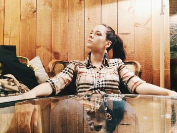 Thoughtful young woman sitting on armchair by table against wooden wall