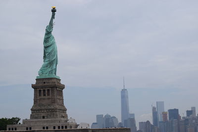 The statue of liberty on a cloudy day in front of new york city and the one world trade center.