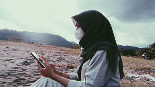 Side view of young woman wearing mask using mobile phone against sky