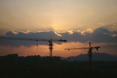 Silhouette cranes by buildings against sky during sunset