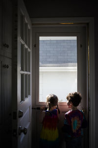 Rear view of siblings looking through window while standing at home