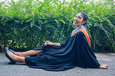 Portrait of smiling young woman in graduation gown sitting against plants