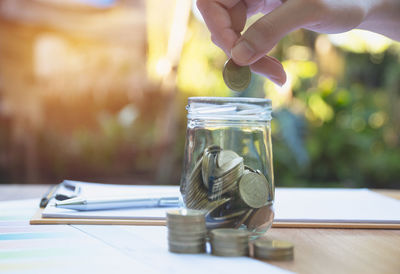 Cropped hand of person holding coin over glass jar by paper and pen on table