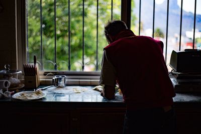 Rear view of man preparing food by window in kitchen