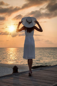 Full length rear view of woman standing by sea against sky during sunset