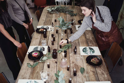 High angle view of mid adult female owner holding perfume bottle on table with colleagues standing at workshop