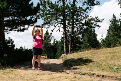 Hiker woman wearing straw hat, shorts and backpack on the path in a forest of pine trees stands up taking a photo of the nature while enjoys the natural environment around. horizontal photo.