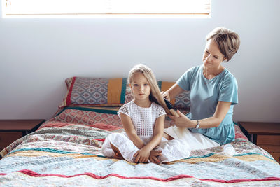 Mother combing daughter hair while sitting on bed at home