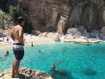 Rear view of shirtless man standing on rocks by sea