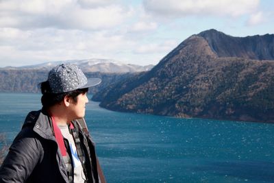 Man standing by river and mountains against cloudy sky