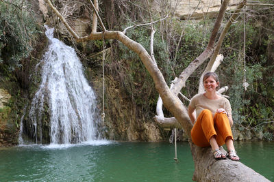 Young woman sitting on tree trunk against waterfall