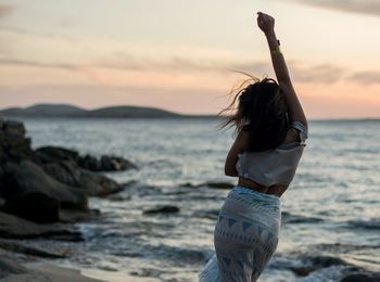 Rear view of woman with arm raised standing at beach against sky during sunset