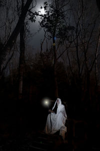 Woman sitting on illuminated street light in forest at night