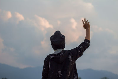 Rear view of man with hand raised standing against cloudy sky