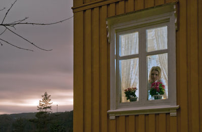 Young woman looking through window of house against sky at dusk