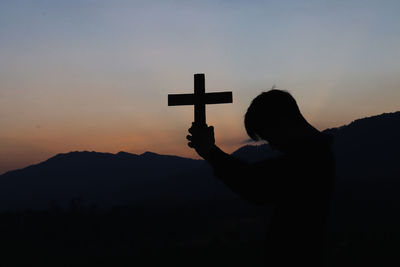 Silhouette person holding cross on mountain against sky during sunset