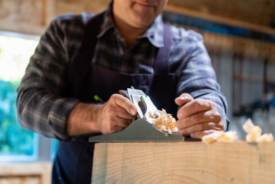 Unrecognizable male woodworker smoothing wooden detail with jack plane while working in professional carpentry workshop